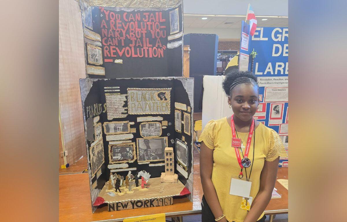 Young girl in a yellow top stands beside a diorama about the Black Panthers and 1969 New York activism at a student exhibit.