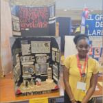 Young girl in a yellow top stands beside a diorama about the Black Panthers and 1969 New York activism at a student exhibit.