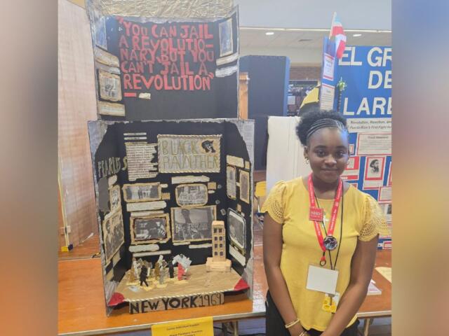 Young girl in a yellow top stands beside a diorama about the Black Panthers and 1969 New York activism at a student exhibit.
