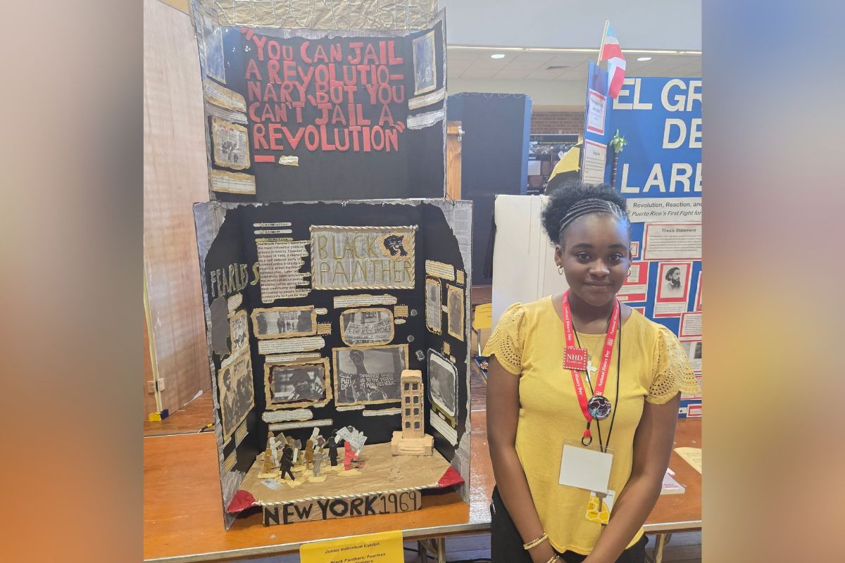 Young girl in a yellow top stands beside a diorama about the Black Panthers and 1969 New York activism at a student exhibit.