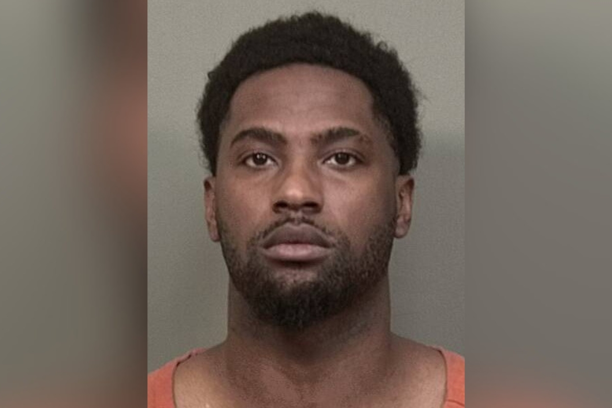 Mugshot of a man with short curly hair and facial hair wearing an orange jail jumpsuit against a grey backdrop.