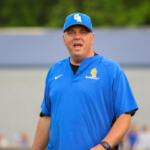 Baseball coach in a blue uniform speaking to players on a field, wearing cap with CA logo and a polo that reads BASEBALL