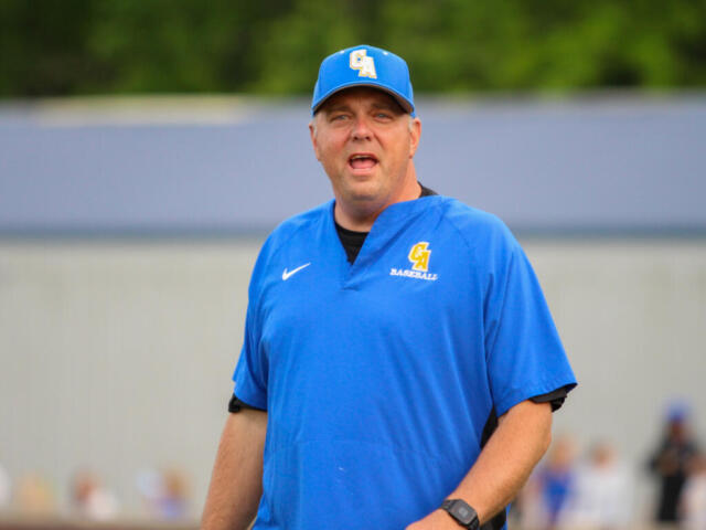 Baseball coach in a blue uniform speaking to players on a field, wearing cap with CA logo and a polo that reads BASEBALL