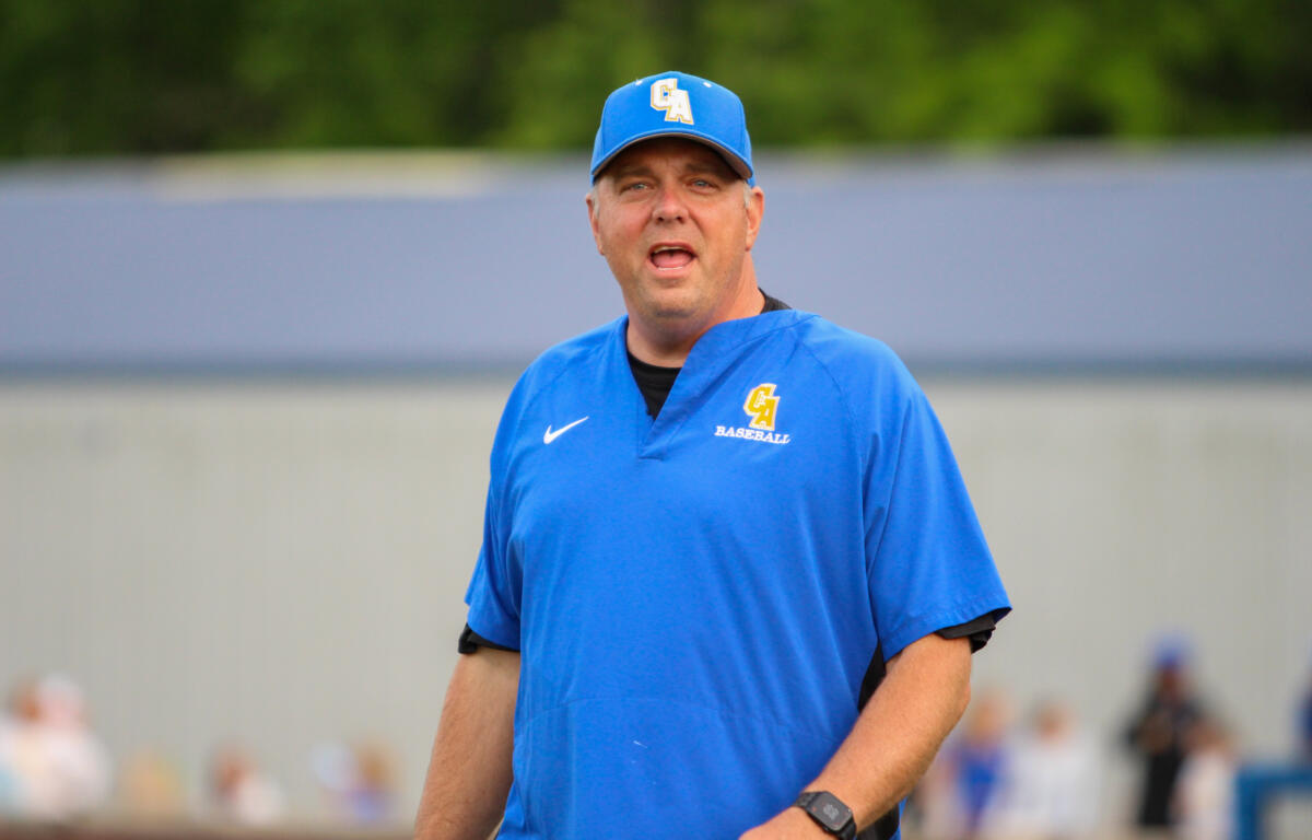 Baseball coach in a blue uniform speaking to players on a field, wearing cap with CA logo and a polo that reads BASEBALL