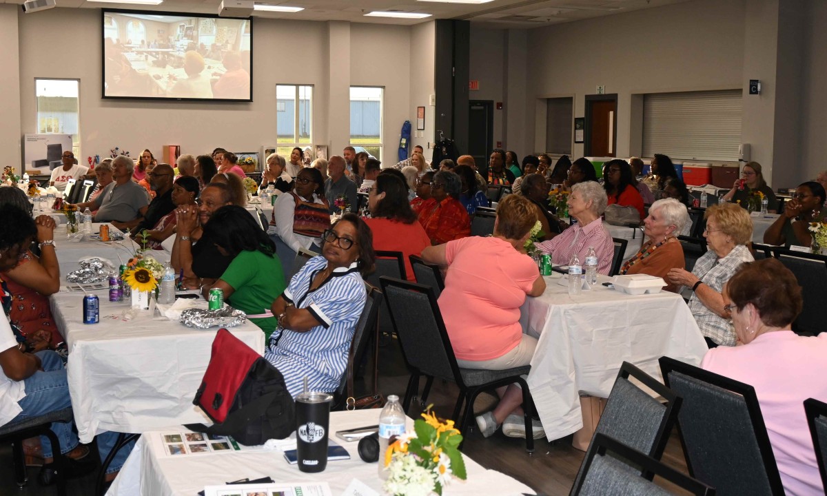 A large group of diverse people seated at round and rectangular tables in a banquet hall, listening to a presentation projected on a screen at the front.