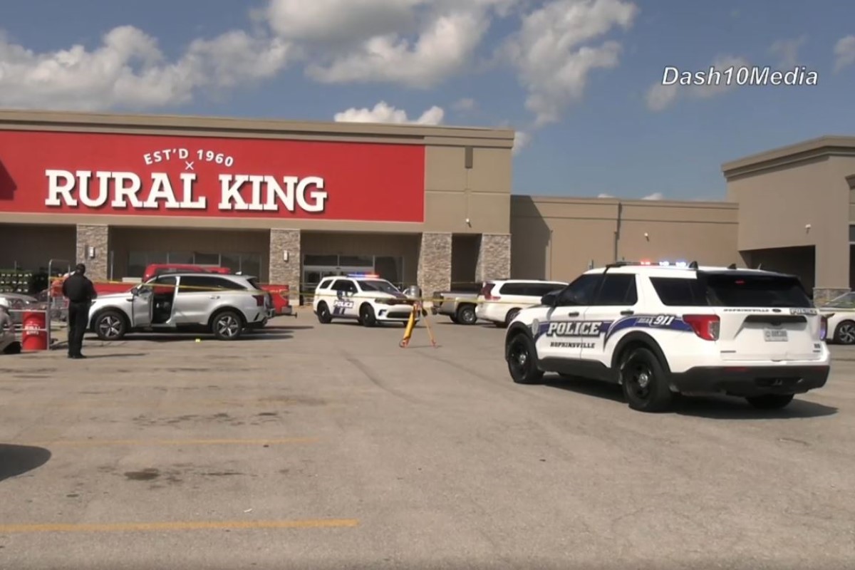 Police cars line the parking lot in front of a Rural King store during an incident, with officers near a white SUV and a barricade in place.