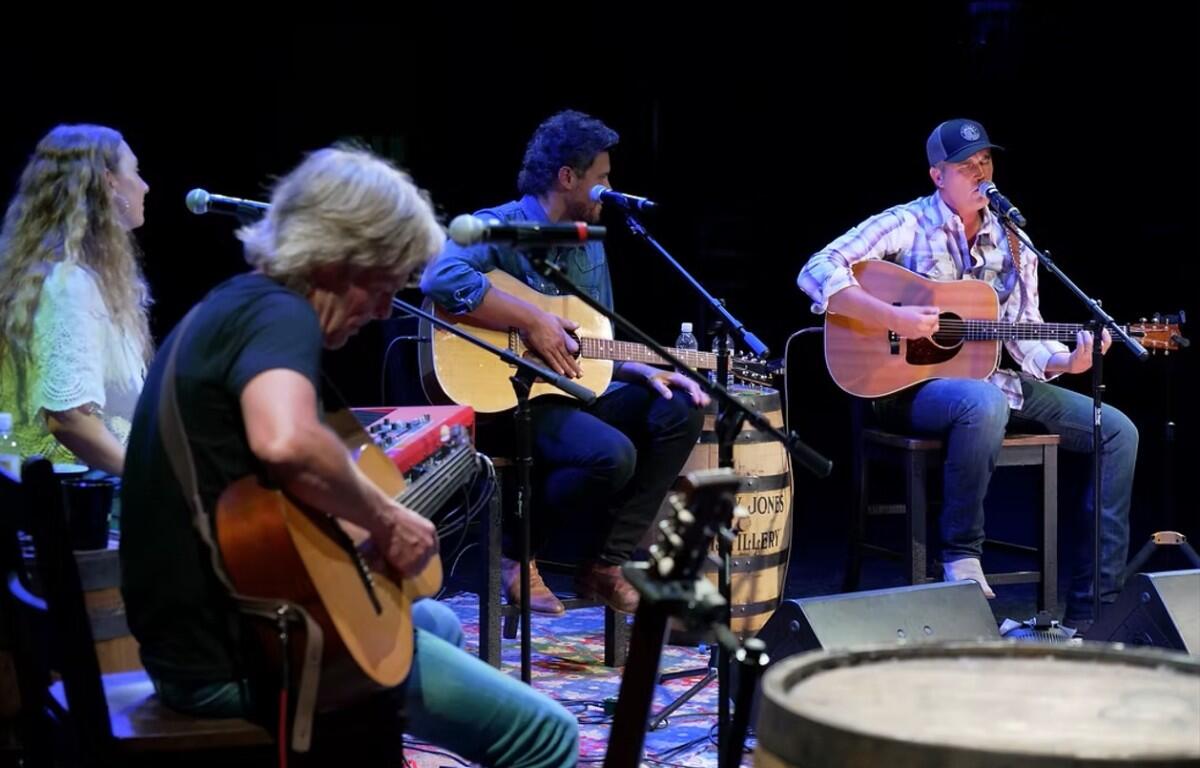 Live acoustic band performing on a dimly lit stage with four guitarists and microphones on stands.