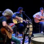 Live acoustic band performing on a dimly lit stage with four guitarists and microphones on stands.