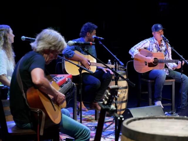 Live acoustic band performing on a dimly lit stage with four guitarists and microphones on stands.