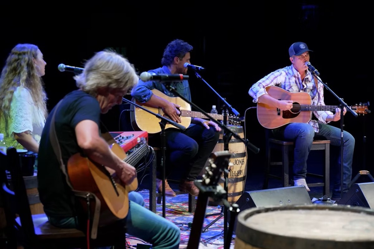 Live acoustic band performing on a dimly lit stage with four guitarists and microphones on stands.