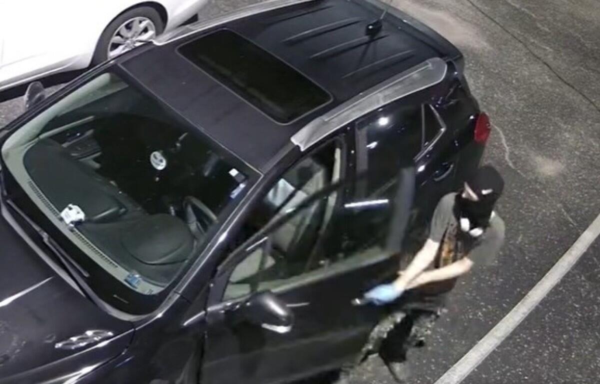 Masked person in a balaclava and gloves standing beside a dark SUV in a parking lot, seen from above (surveillance-style).