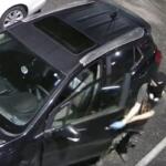 Masked person in a balaclava and gloves standing beside a dark SUV in a parking lot, seen from above (surveillance-style).