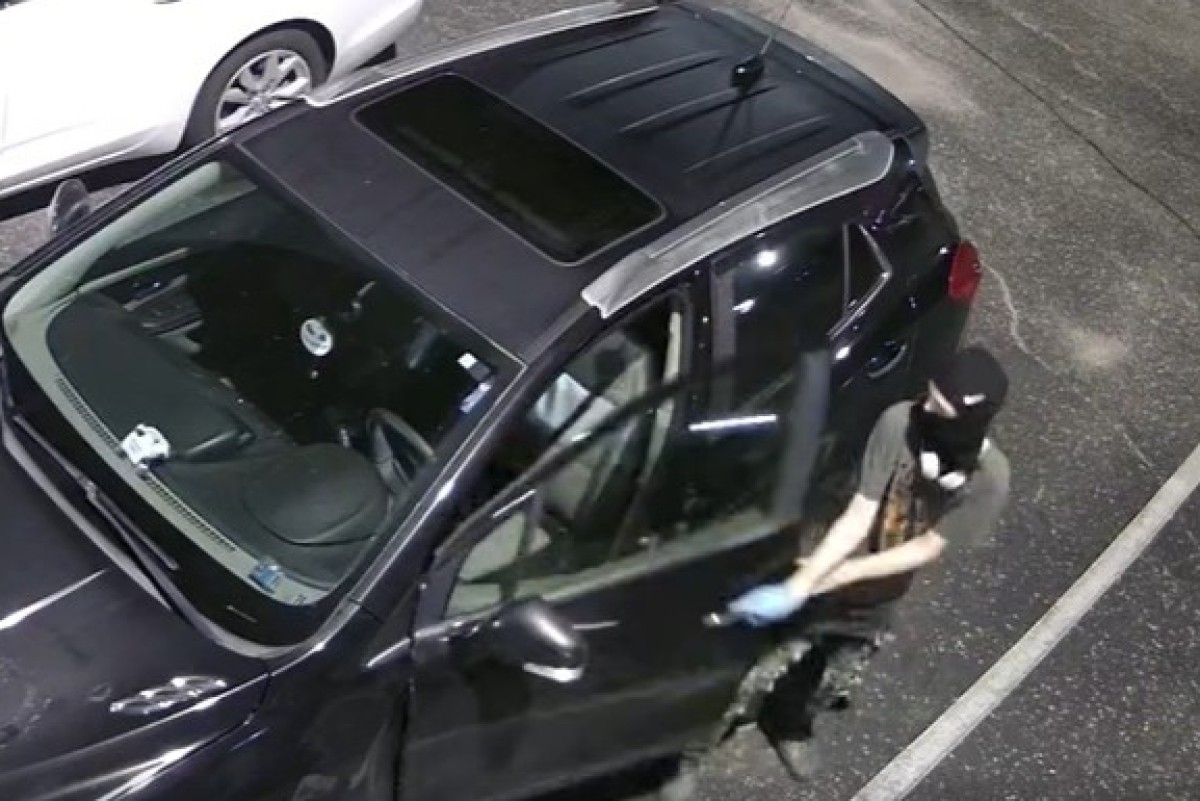 Masked person in a balaclava and gloves standing beside a dark SUV in a parking lot, seen from above (surveillance-style).