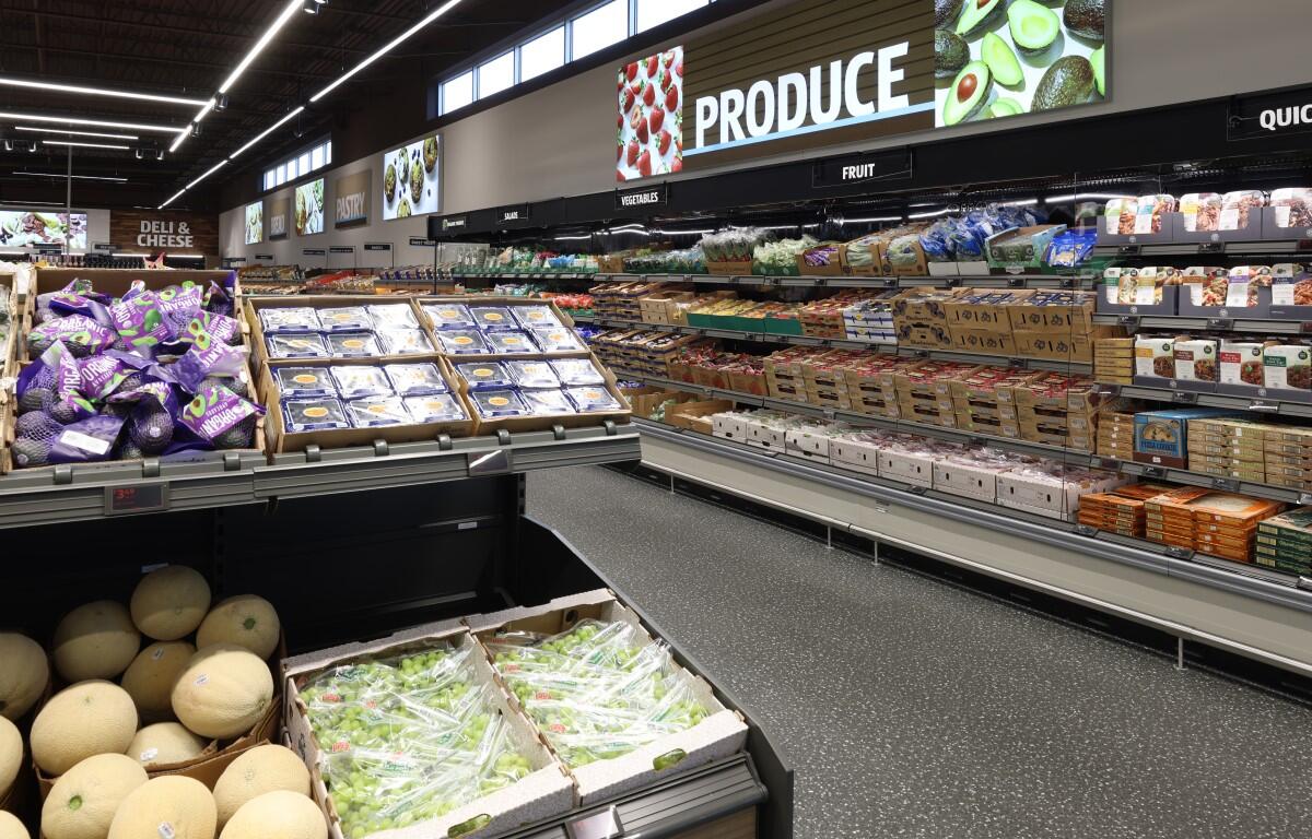 A bright grocery store produce section with shelves of fruits, vegetables, and packaged goods along a long aisle.
