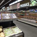 A bright grocery store produce section with shelves of fruits, vegetables, and packaged goods along a long aisle.