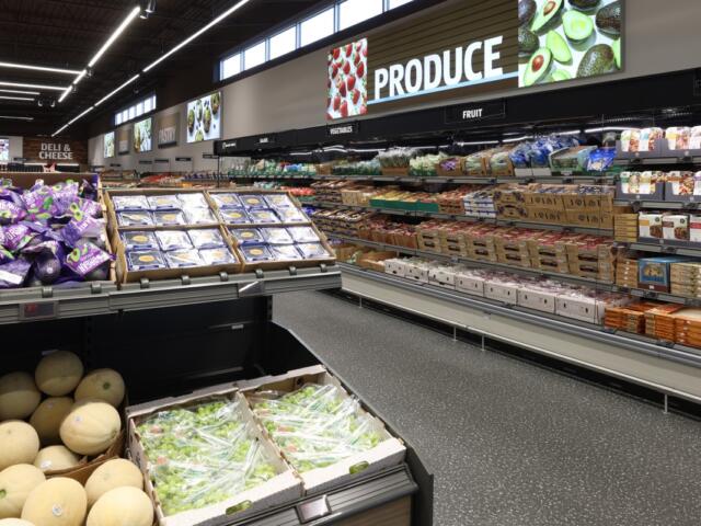 A bright grocery store produce section with shelves of fruits, vegetables, and packaged goods along a long aisle.