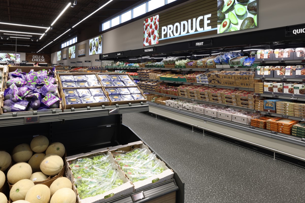 A bright grocery store produce section with shelves of fruits, vegetables, and packaged goods along a long aisle.