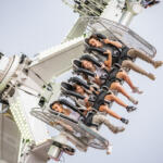 Six riders strapped into seats on a high, rotating amusement ride against a clear sky, expressions of excitement on their faces.