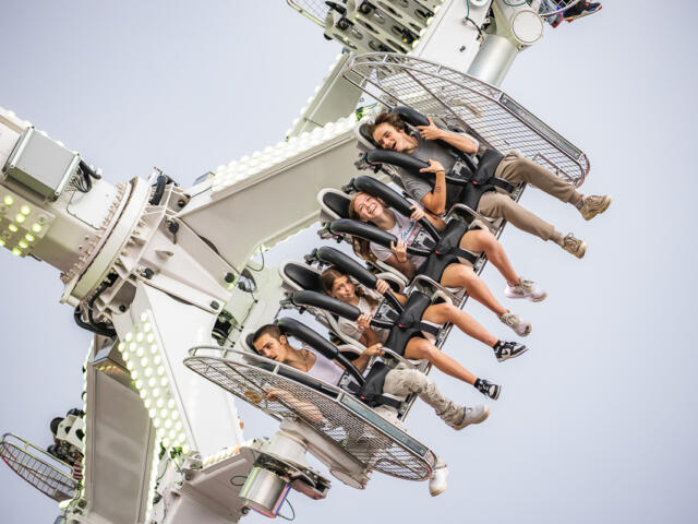 Six riders strapped into seats on a high, rotating amusement ride against a clear sky, expressions of excitement on their faces.