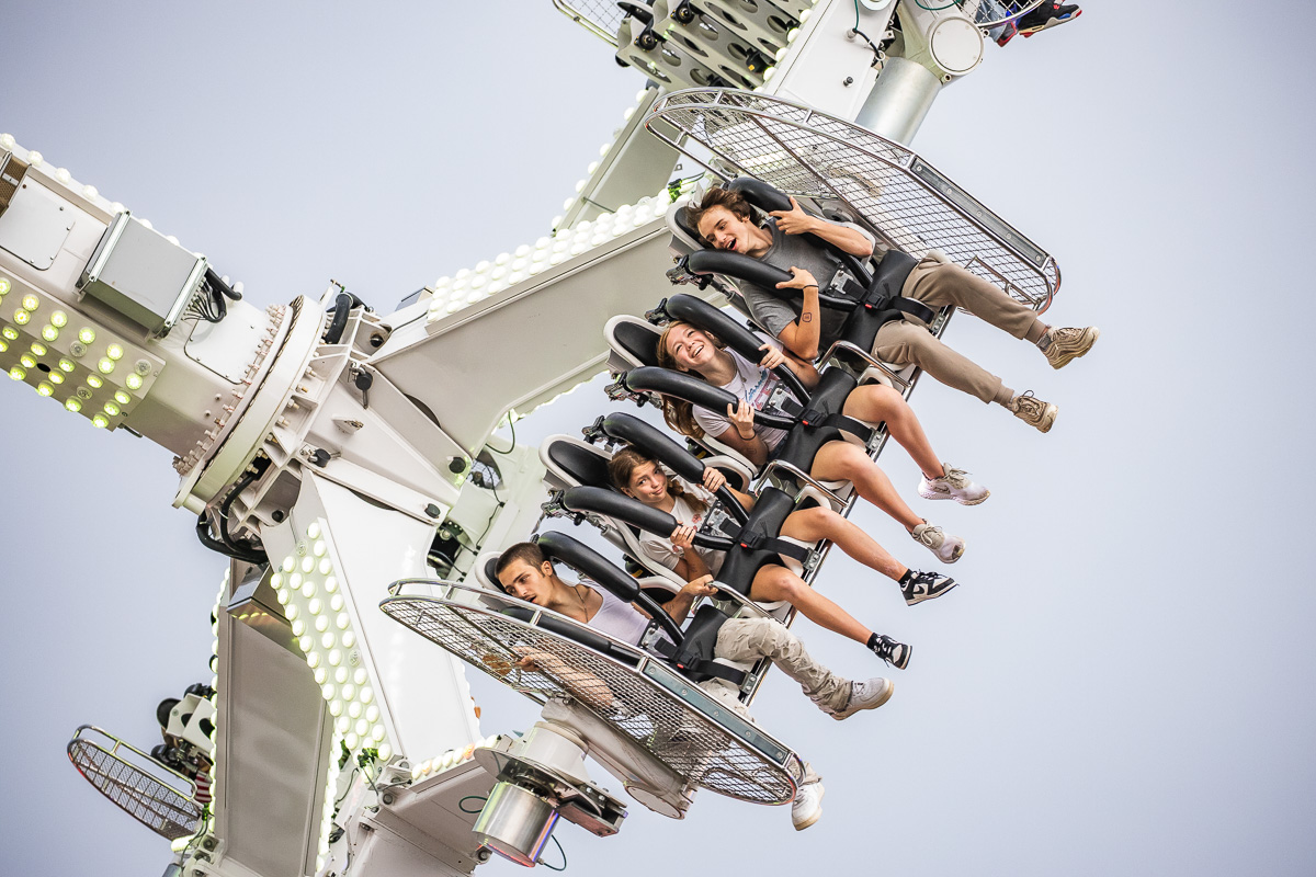 Six riders strapped into seats on a high, rotating amusement ride against a clear sky, expressions of excitement on their faces.