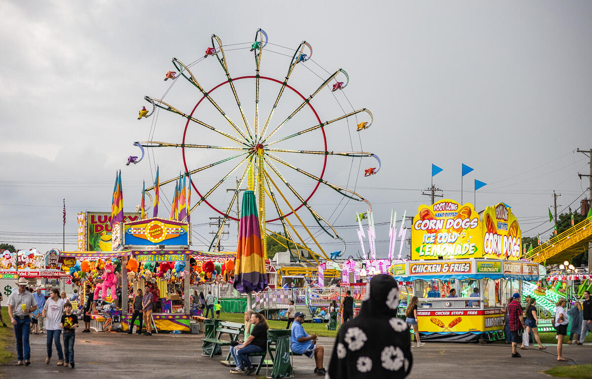Colorful carnival scene with a large illuminated Ferris wheel behind game stalls and food stands.