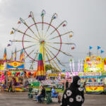 Colorful carnival scene with a large illuminated Ferris wheel behind game stalls and food stands.