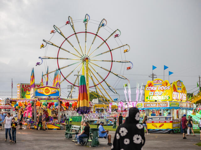 Colorful carnival scene with a large illuminated Ferris wheel behind game stalls and food stands.