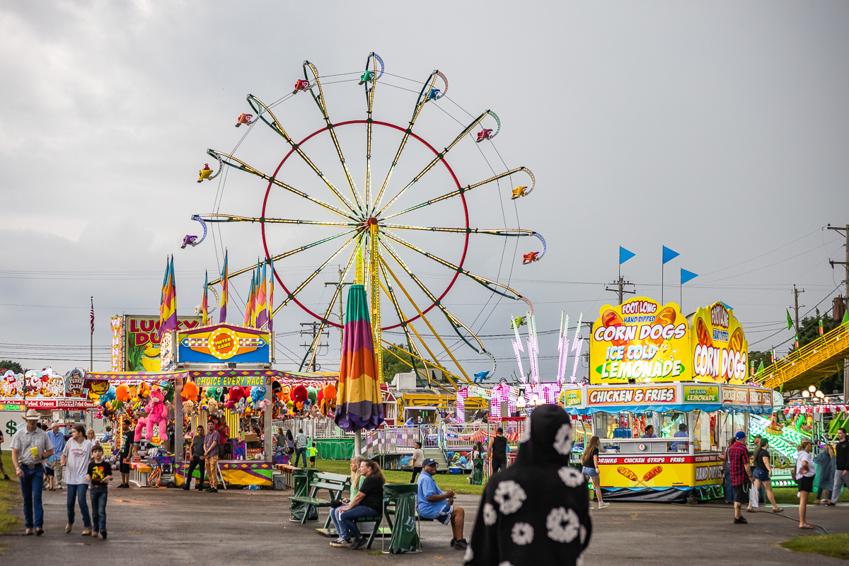 Colorful carnival scene with a large illuminated Ferris wheel behind game stalls and food stands.