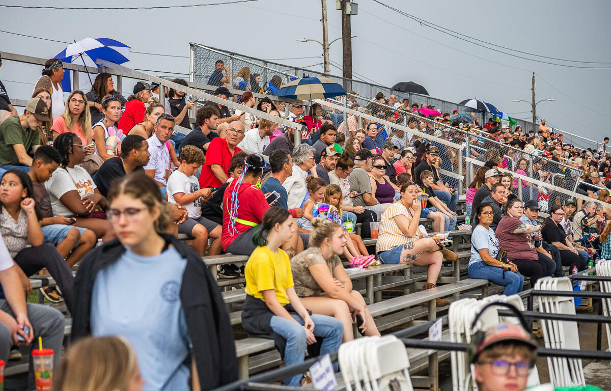 Crowded outdoor bleachers filled with diverse spectators watching an event, some people holding drinks and umbrellas against a sunny day.