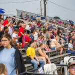 Crowded outdoor bleachers filled with diverse spectators watching an event, some people holding drinks and umbrellas against a sunny day.