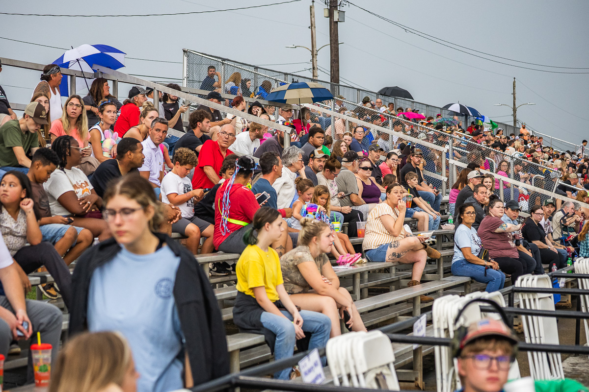 Crowded outdoor bleachers filled with diverse spectators watching an event, some people holding drinks and umbrellas against a sunny day.