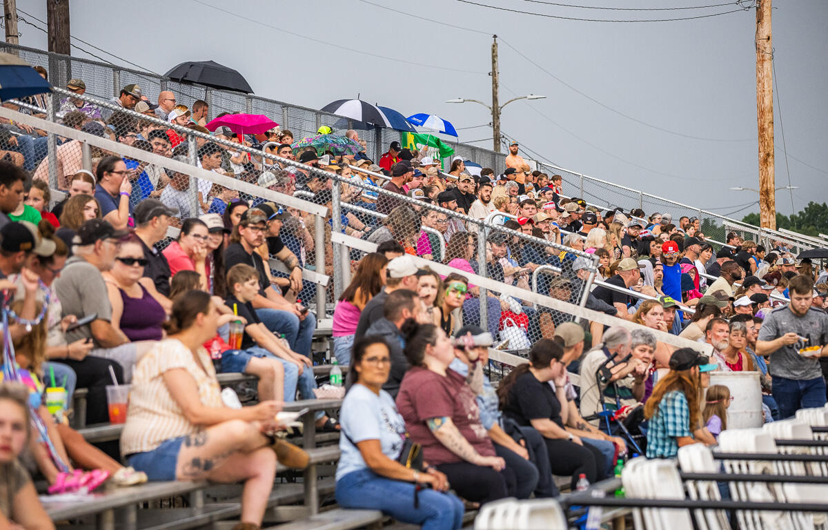 Crowd of people sitting on metal stadium bleachers at an outdoor event, some with umbrellas and drinks, behind a chain-link fence.