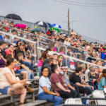 Crowd of people sitting on metal stadium bleachers at an outdoor event, some with umbrellas and drinks, behind a chain-link fence.