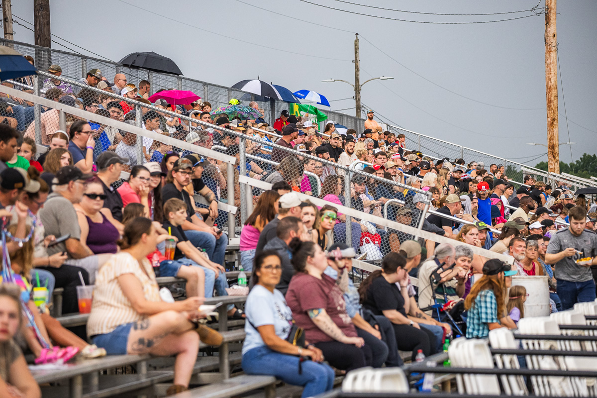 Crowd of people sitting on metal stadium bleachers at an outdoor event, some with umbrellas and drinks, behind a chain-link fence.