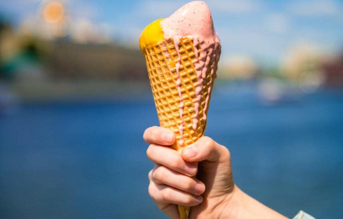 Hand holding a waffle cone with pink and yellow ice cream against a waterfront cityscape as background.