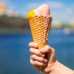 Hand holding a waffle cone with pink and yellow ice cream against a waterfront cityscape as background.
