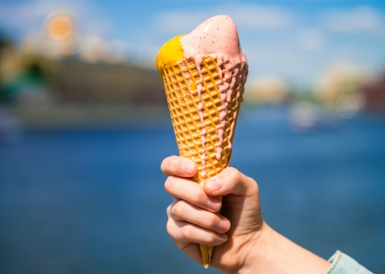 Hand holding a waffle cone with pink and yellow ice cream against a waterfront cityscape as background.