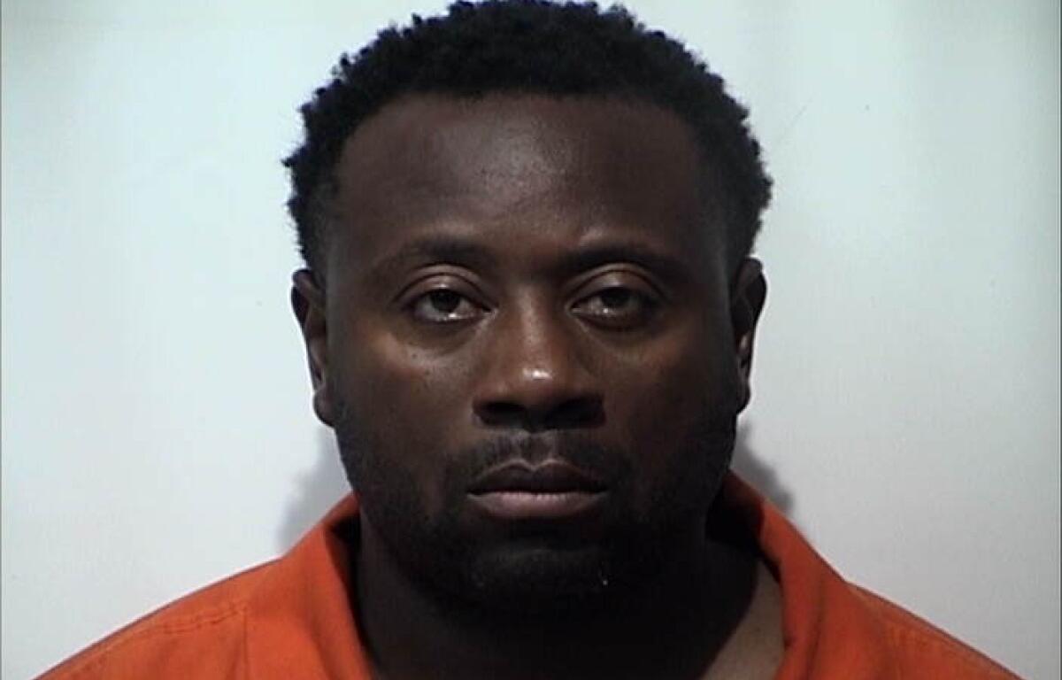 Mugshot of a Black man with short curly hair, wearing an orange prison jumpsuit, facing the camera against a plain light background.
