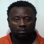 Mugshot of a Black man with short curly hair, wearing an orange prison jumpsuit, facing the camera against a plain light background.