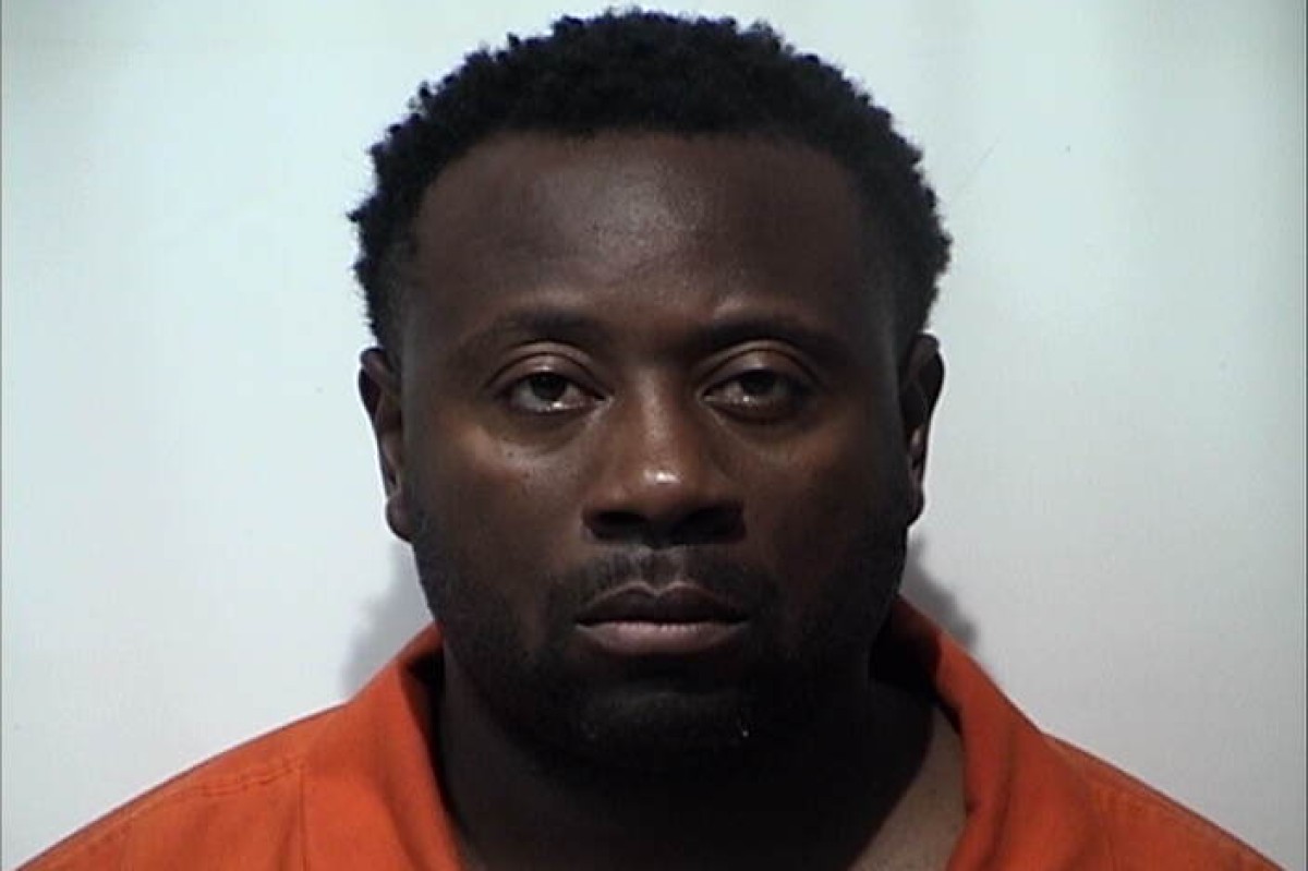 Mugshot of a Black man with short curly hair, wearing an orange prison jumpsuit, facing the camera against a plain light background.