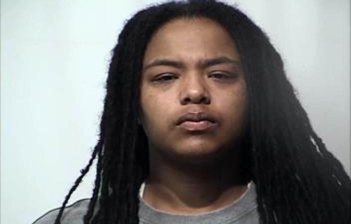 Mugshot-style portrait of a young person with long black dreadlocks and a gray shirt, neutral expression.