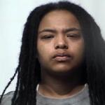 Mugshot-style portrait of a young person with long black dreadlocks and a gray shirt, neutral expression.