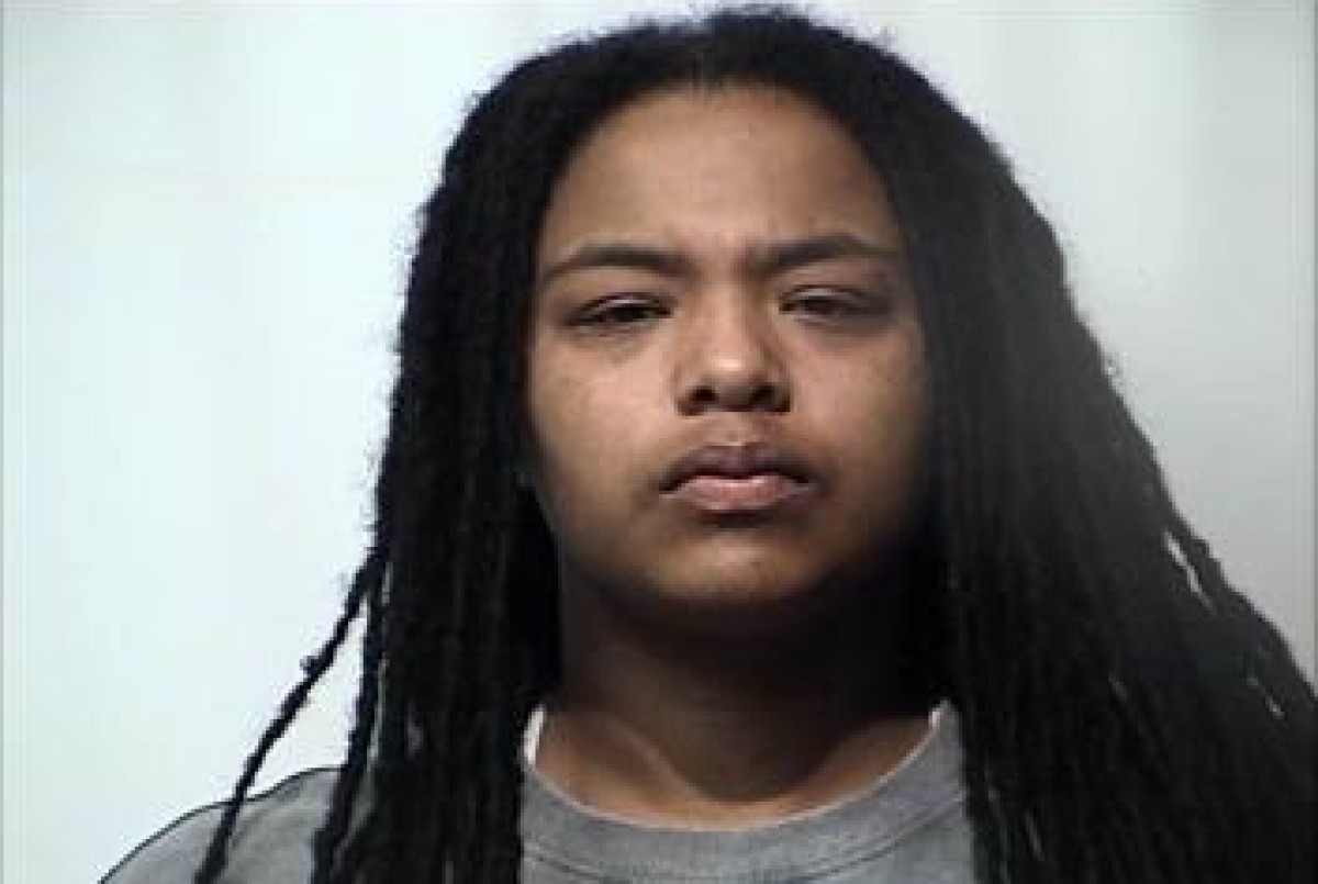 Mugshot-style portrait of a young person with long black dreadlocks and a gray shirt, neutral expression.