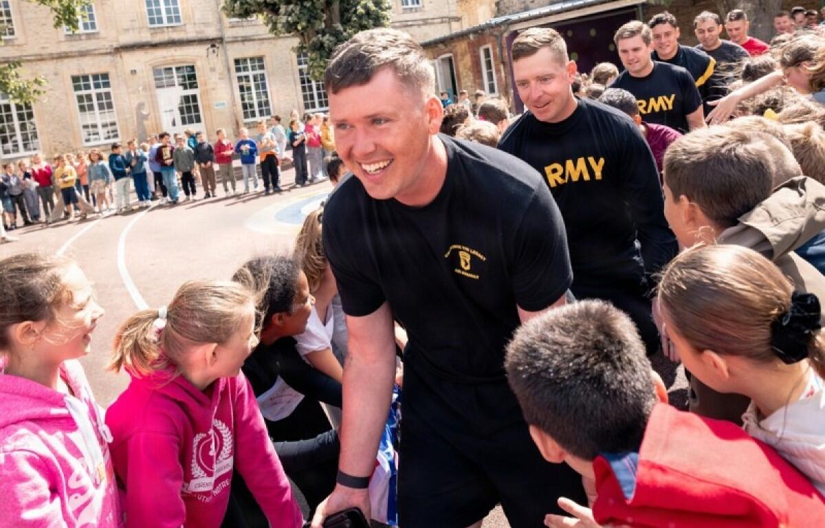 Soldier in black athletic gear high-fives smiling children in a schoolyard as a line of spectators watches from the background.