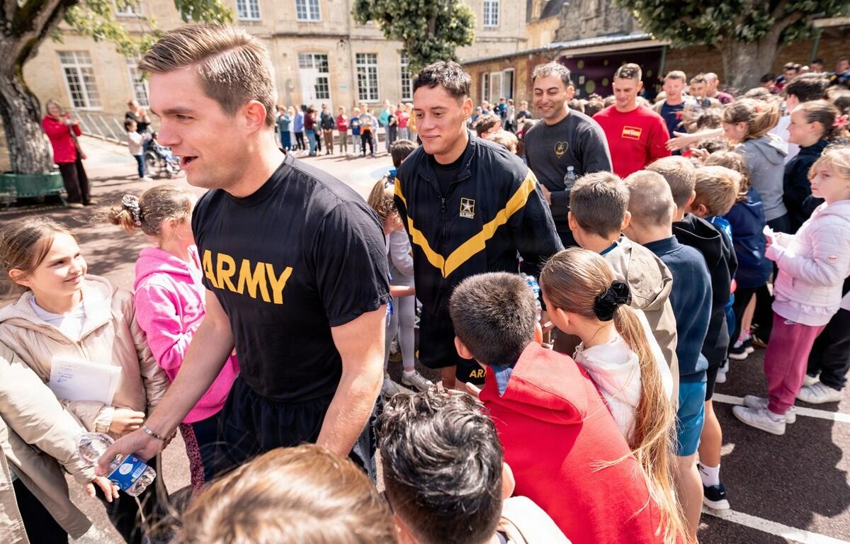 Soldier in a black Army shirt shakes hands with a smiling girl at a crowded outdoor community event in a schoolyard setting.