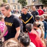 Soldier in a black Army shirt shakes hands with a smiling girl at a crowded outdoor community event in a schoolyard setting.