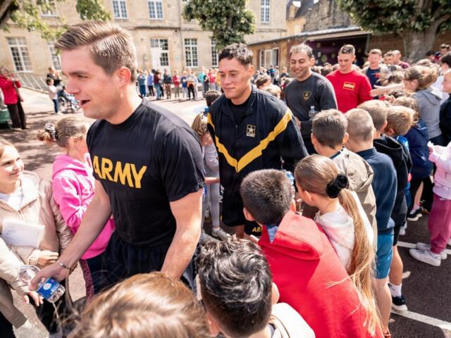 Soldier in a black Army shirt shakes hands with a smiling girl at a crowded outdoor community event in a schoolyard setting.