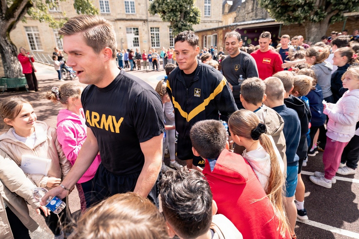Soldier in a black Army shirt shakes hands with a smiling girl at a crowded outdoor community event in a schoolyard setting.