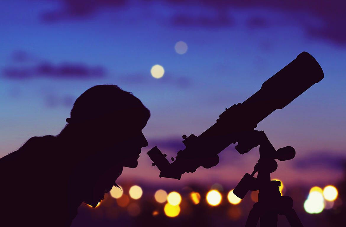 Silhouetted person looking through a telescope at dusk with city lights in the background