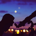 Silhouetted person looking through a telescope at dusk with city lights in the background